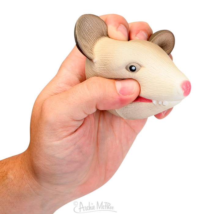 A hand squeezing the Stress Possum toy, crafted from soft polyurethane foam and designed to resemble a beige possum head with a pink nose and brown ears, offers calming stress relief.