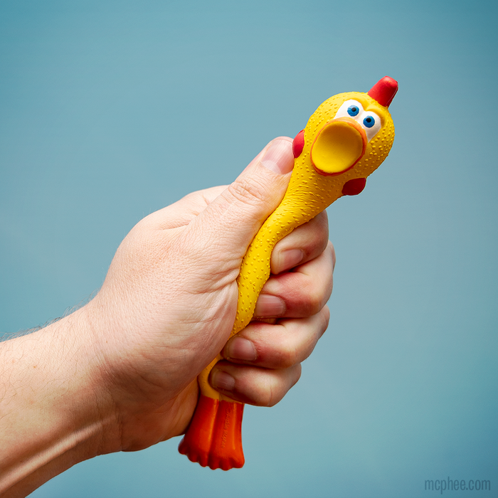 Against a blue background, a hand squeezes the Stress Rubber Chicken toy made from squishy polyurethane foam for stress relief.