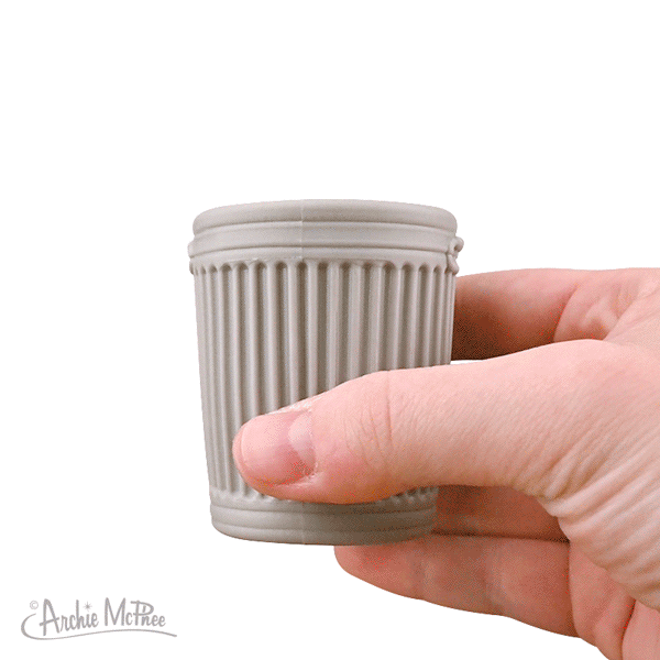 A hand holds a gray coffee cup, known as the Surprise Popping Alley Cat, shaped like a trash can, with the lid magically transforming.