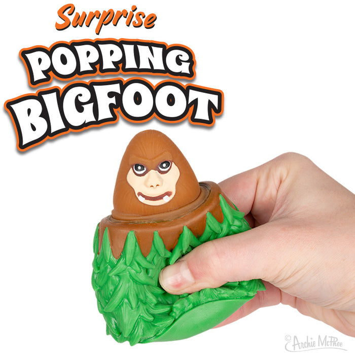 A hand presses a rubber toy, causing a cartoon Bigfoot face to pop out from a green leafy base. The text states the product name: Surprise Popping Bigfoot.