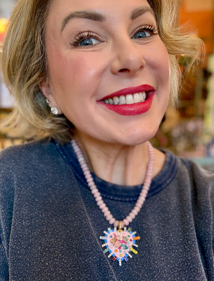 A Tova brand beaded necklace featuring light pink faceted beads, a gold-tone clasp, and a large, colorful heart-shaped pendant with cartoon-style illustrations and rhinestones.