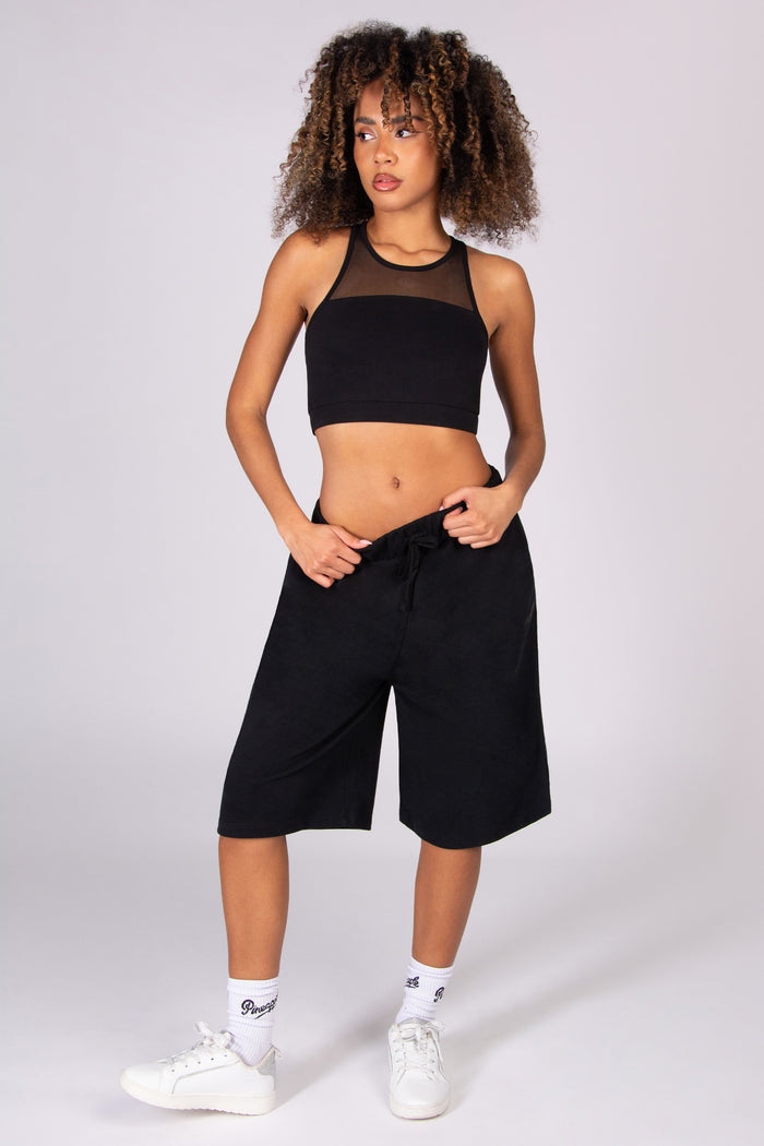 Woman wearing a black crop top and black super oversized basketball shorts  on a light grey background