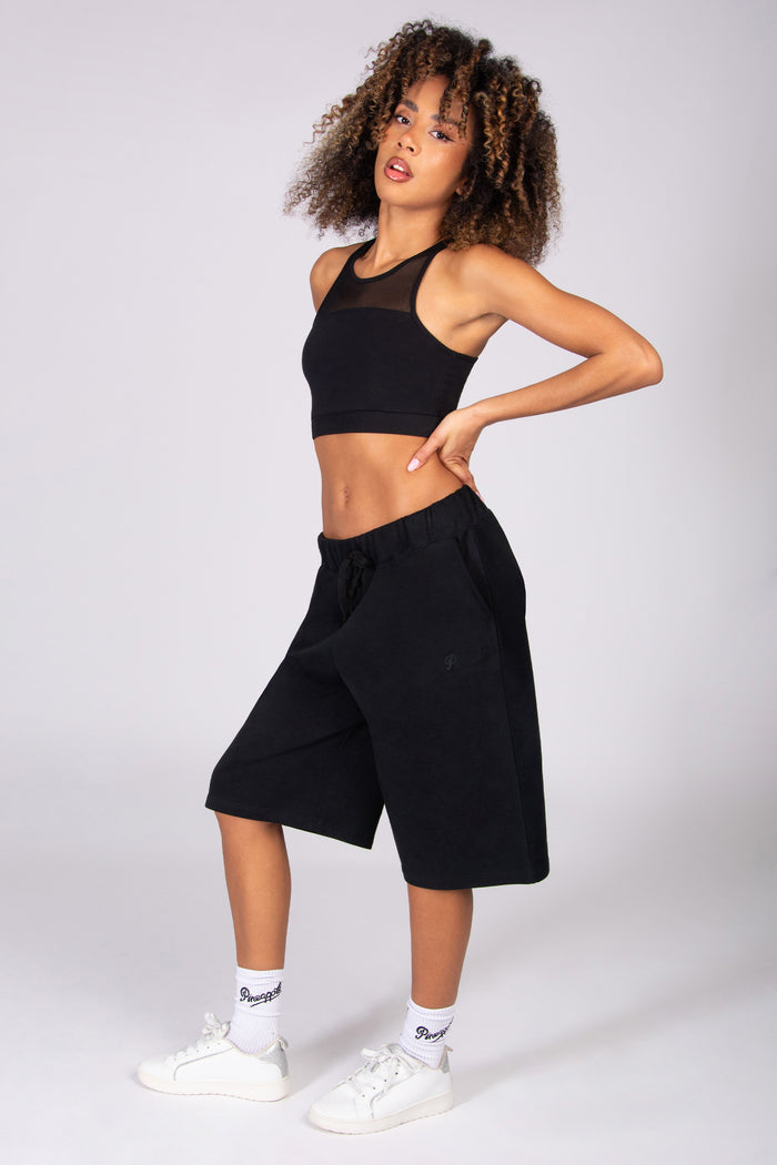 Woman wearing a black crop top and black super oversized basketball shorts  on a light grey background