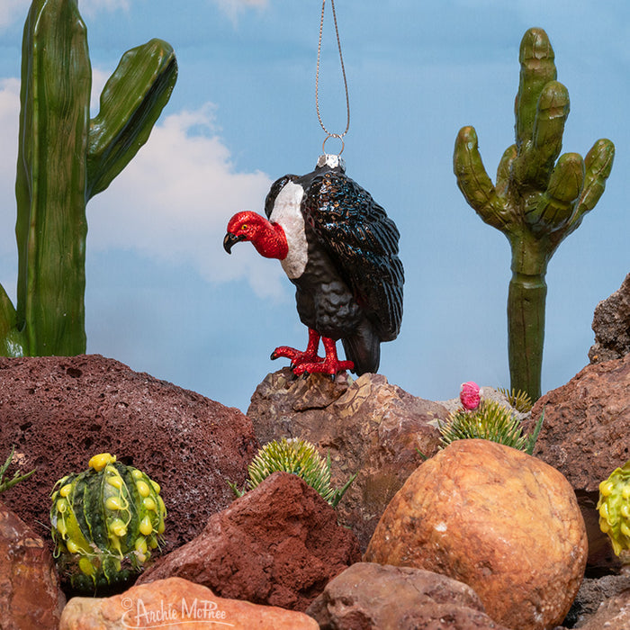 The Vulture Ornament rests on a rocky landscape with cacti against a clear blue sky, showcasing the stark beauty of the desert environment.