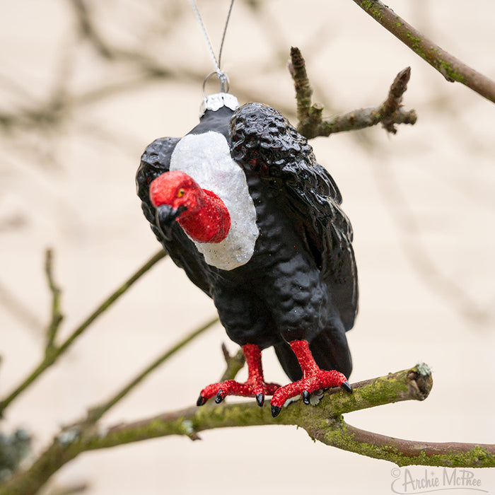 The Vulture Ornament, with its hand-blown glass design, striking red head and feet, hangs elegantly from a tree branch. This unique piece makes for a distinctive Christmas decoration, capturing attention with its intricate detail and craftsmanship.