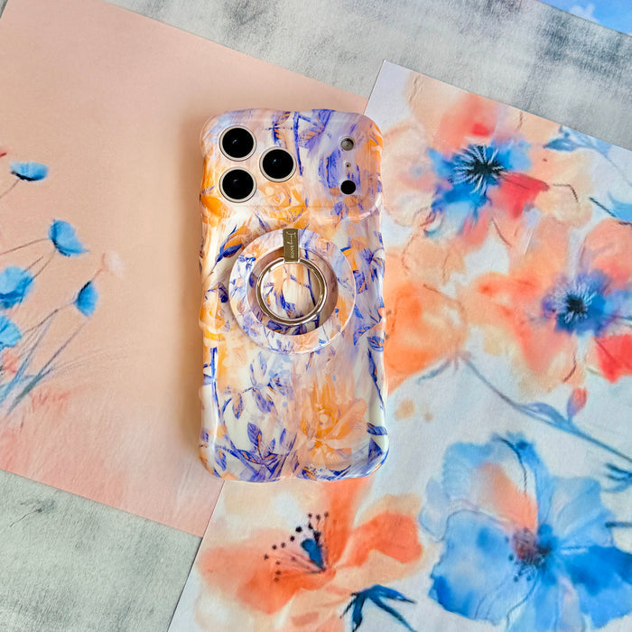 Where Dreams Bloom Wavy Luxe iPhone case with the matching MagSafe grip ring attached, styled on coral and blue watercolor floral paper.