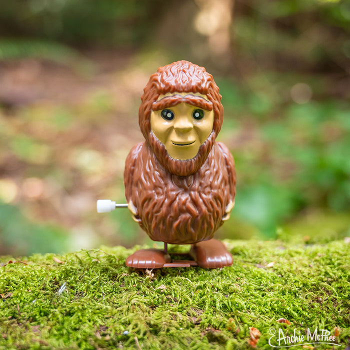 A Wind-Up Bigfoot, featuring a vinyl design and a wind-up key on its side, stands amidst moss in a forest scene, making it perfect for an illustrated display box.