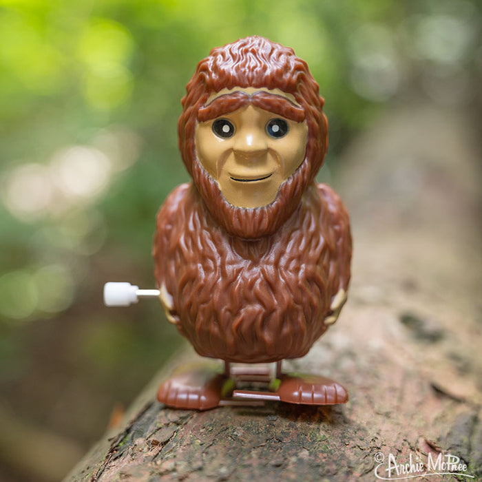 The Wind-Up Bigfoot, a vinyl toy with a cheerful expression, stands proudly on a log in a forest setting.